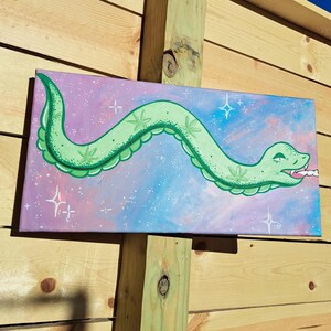 May include: A green snake with a leafy pattern is painted on a canvas with a pink, blue, and white galaxy background. The snake is holding a white cigarette in its mouth.