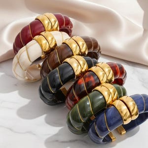 May include: A collection of colorful, chunky bangles. The bracelets are in various colors, including red, cream, brown, black, tortoiseshell, green, and blue. Each bangle features a gold-tone metal accent. The bracelets are arranged on a white surface.
