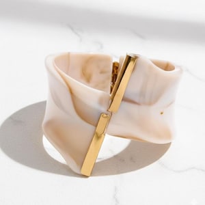 May include: A wide, cream-colored cuff bracelet with a marbled effect and a gold-colored clasp. The bracelet has a unique, sculptural design and is made of a smooth, polished material. The gold clasp is rectangular and connects the two sides of the bracelet.