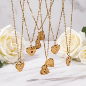 May include: Gold-colored heart-shaped lockets on delicate chains. Some lockets are closed, while others are open, revealing a small space inside. Each locket features a small gemstone. The background includes white roses.