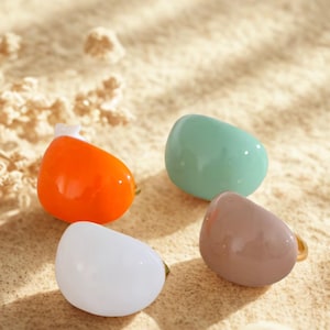 May include: Four colorful, smooth, teardrop-shaped rings. The rings are in shades of orange, mint green, white, and taupe. Each ring has a gold-colored band. The rings are displayed on a sandy surface.