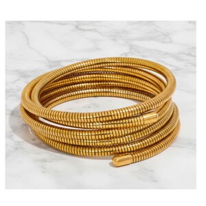 May include: A gold-colored, coiled bracelet with a snake-like design. The bracelet is made of multiple segments, creating a flexible and textured appearance. The ends of the bracelet have a smooth, rounded finish. The bracelet is displayed on a white marble surface.