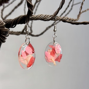 Pu&ograve; includere: Orecchini ovali con resina trasparente, che mostrano petali di fiori rosa e rossi pressati. Gli orecchini pendono da ganci per orecchie color argento. Lo sfondo &egrave; un grigio neutro sfocato.