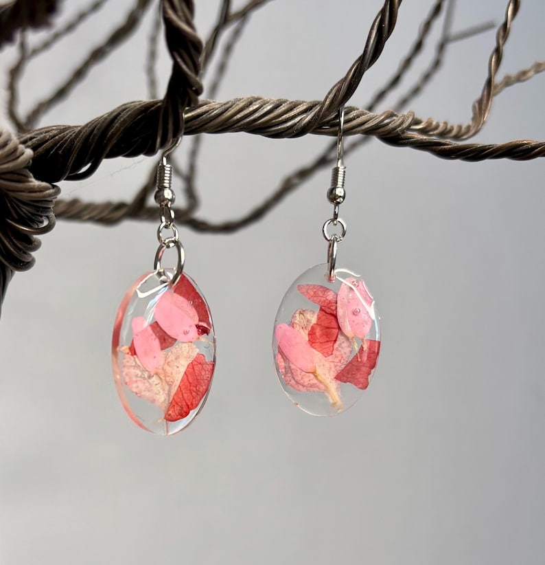 Pu&ograve; includere: Orecchini ovali con resina trasparente, che mostrano petali di fiori rosa e rossi pressati. Gli orecchini pendono da ganci per orecchie color argento. Lo sfondo &egrave; un grigio neutro sfocato.