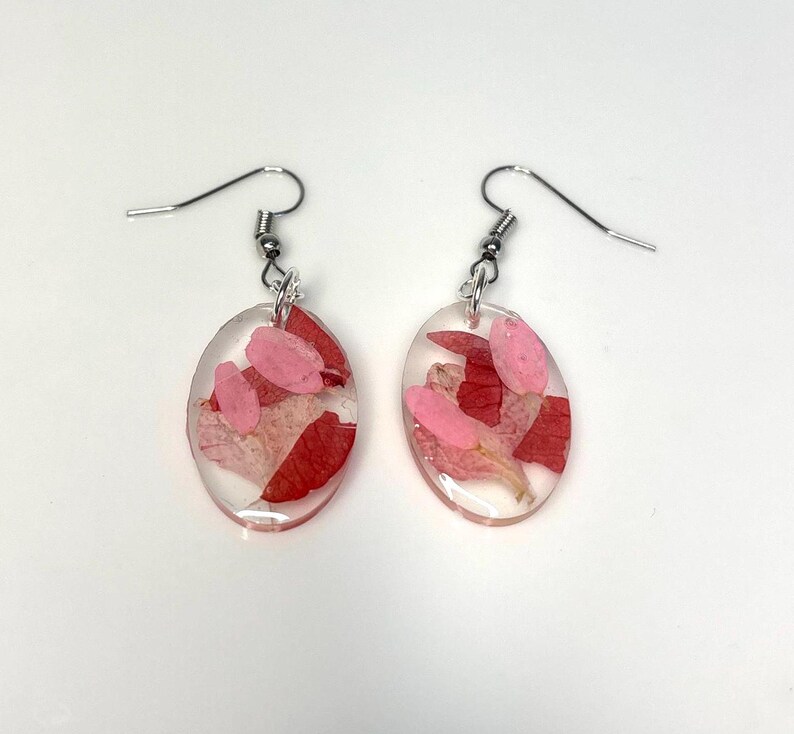 Pu&ograve; includere: Un paio di orecchini ovali con ganci argentati. Ogni orecchino &egrave; riempito di resina trasparente e contiene petali di fiori rosa e rossi. Gli orecchini sono esposti su uno sfondo bianco.
