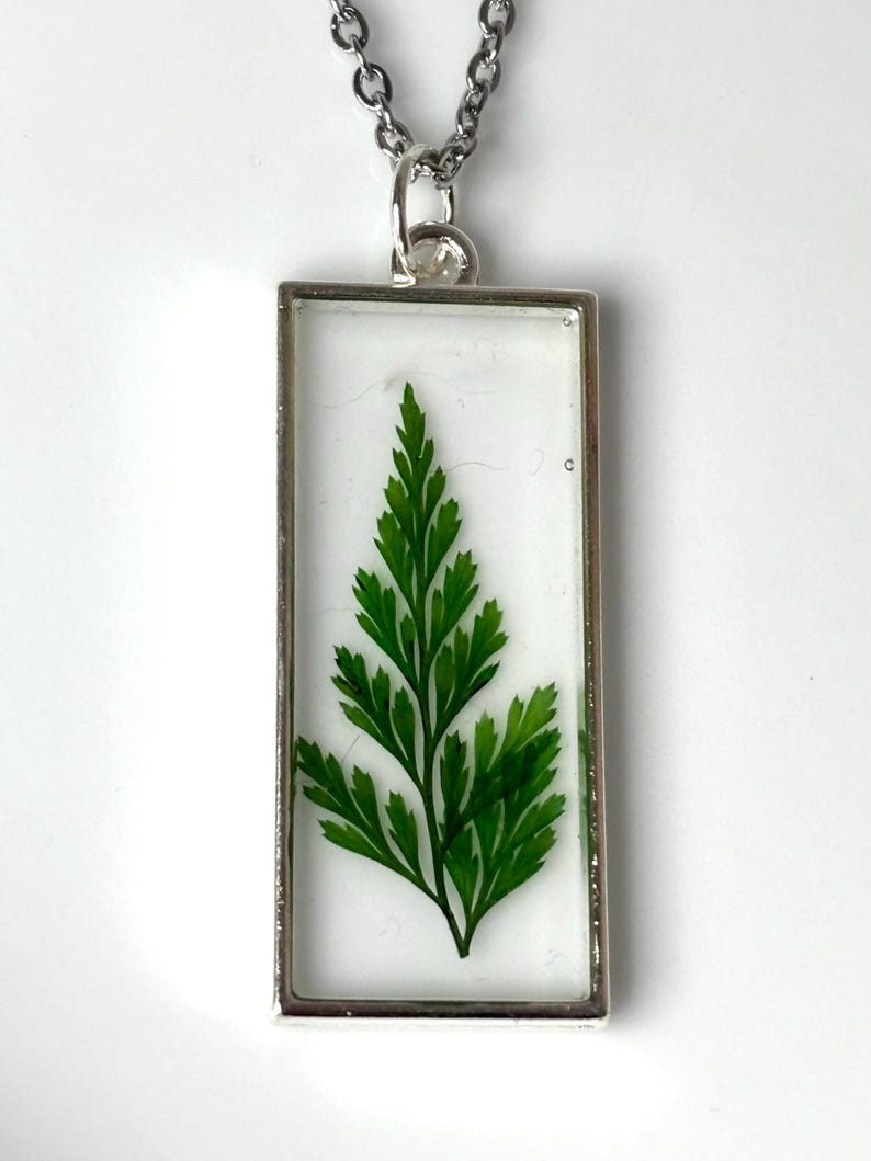 Pu&ograve; includere: Una collana con pendente rettangolare in argento con una vivace foglia di felce verde racchiusa in resina trasparente. I dettagli intricati della foglia sono visibili, su uno sfondo bianco. Il pendente &egrave; appeso a una delicata catena d'argento.