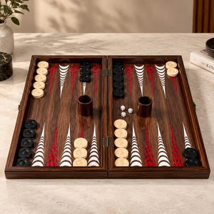May include: A wooden backgammon board featuring a dark brown wood grain finish with alternating red and white triangular points. The set includes light beige and black circular checkers, two dark brown dice cups, and two small white dice with black dots.