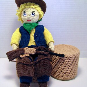 Pode incluir: Boneca de crochê feita à mão vestida de cowboy, usando chapéu marrom, colete azul, camisa amarela e calças marrons. A boneca tem cabelo loiro, olhos azuis e um lenço verde. Um banquinho de crochê correspondente está ao lado.