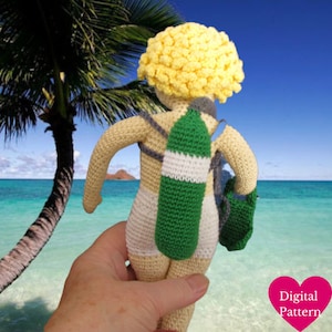 Op de afbeelding: Gehaakte amigurumi pop met geel haar, draagt een witte badpak en draagt een groene en witte duikfles op haar rug. De pop houdt een groene en witte duikvin in haar rechterhand. De pop staat op een hand voor een strandscène.