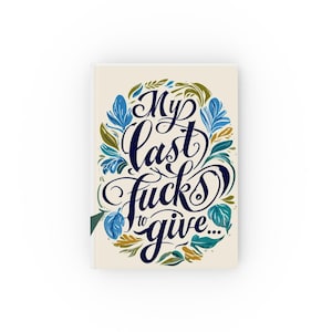 May include: A hardcover journal with the text "My last fucks to give..." in elegant script. The text is surrounded by a wreath of blue, green, and gold leaves. The journal has a cream-colored cover.