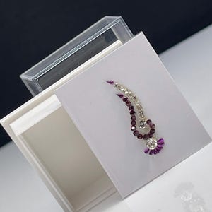 May include: A white box with a clear lid, containing a single purple and white jeweled bindi sticker. The bindi is shaped like a teardrop with a fan-shaped bottom.