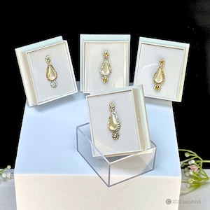 May include: Four white boxes with clear lids display a set of four nose rings. Each ring is a teardrop shape with a white pearl centre and clear rhinestones around the pearl. The rings are displayed on a white surface.