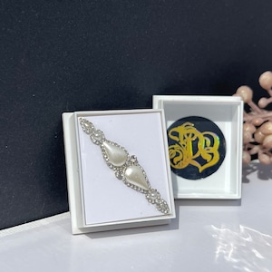 May include: A white box containing a white pearl and rhinestone design. The box is open and the design is visible. The box is on a black surface.