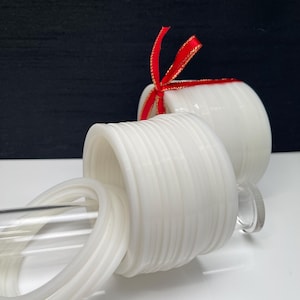 May include: A collection of white, circular, ribbed bangles stacked on a clear acrylic rod. One stack is tied with a thin red ribbon with gold edges. The bangles have a smooth, uniform finish and a consistent, repeating ridged texture throughout.