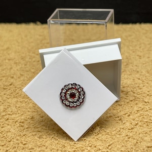 May include: A small white box with a clear lid, containing a red and white jeweled round ornament.