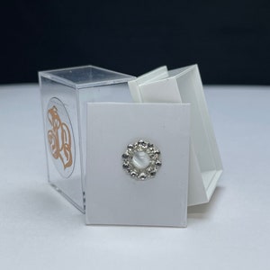 May include: A small, white, round, decorative piece with a silver border and a clear centre. The piece is in a white box with a clear plastic lid.