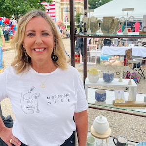 May include: A woman wearing a white t-shirt with the text "Mississippi Made" stands in front of a table displaying handmade pottery and jewelry. The pottery includes mugs, bowls, and a sugar bowl. The jewelry includes earrings and a necklace.