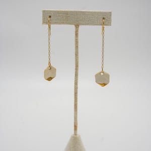 May include: A pair of gold-toned chain earrings with hexagonal, off-white, and gold-edged pendants. The earrings are displayed on a neutral-toned earring stand against a white background. The earrings are a simple, elegant design.