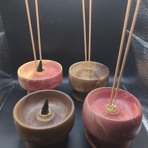 May include: Four wooden incense holders in shades of brown and pink, each with a brass incense holder and a lit incense cone. Incense sticks are placed in the holders. The holders are on a dark surface.