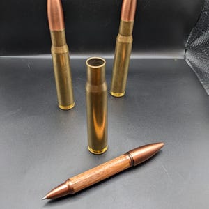 May include: A handmade pen with a wooden barrel and copper accents, alongside three brass bullet casings and a single spent casing. The pen features a copper tip and end, with a polished wood grain finish.