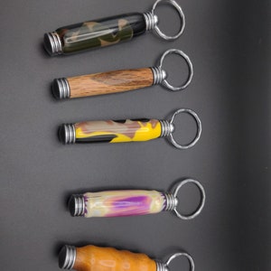 May include: Five different keychains with wooden and resin handles. The keychains are all attached to silver key rings. The handles are in various colors and patterns, including camouflage, wood grain, yellow and brown, purple and white, and brown.