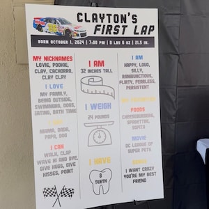 May include: A white sign on a black easel with the text "Clayton's First Lap." The sign includes details about the child's birth, nicknames, height (32 inches), weight (24 pounds), and favorite things. A race car graphic is at the top.