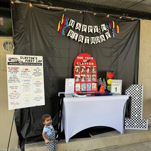 May include: A first birthday party setup with a racing theme. A banner reads "Happy 1st Birthday." Decorations include a checkered flag, a speed limit sign, and a large number one. A child in checkered overalls stands in front of the decorations.