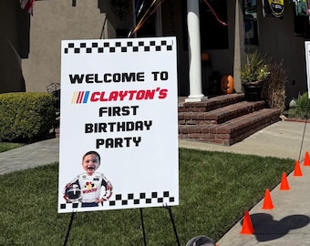 May include: A sign that reads "WELCOME TO CLAYTON'S FIRST BIRTHDAY PARTY" with a photo of a baby in a racing suit. The sign is on an easel in front of a house, with balloons and an American flag in the background.