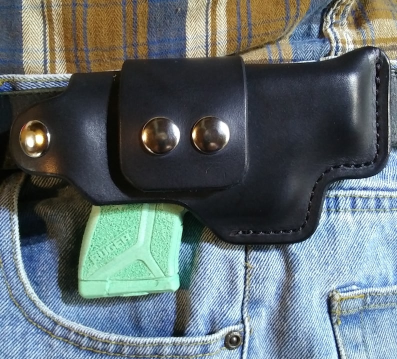 以下が含まれることがあります： シルバー色の金属リベットが付いた黒革ホルスター。緑色の拳銃を保持しています。ホルスターはベルトに取り付けられ、青いデニムジーンズのポケットに差し込まれています。