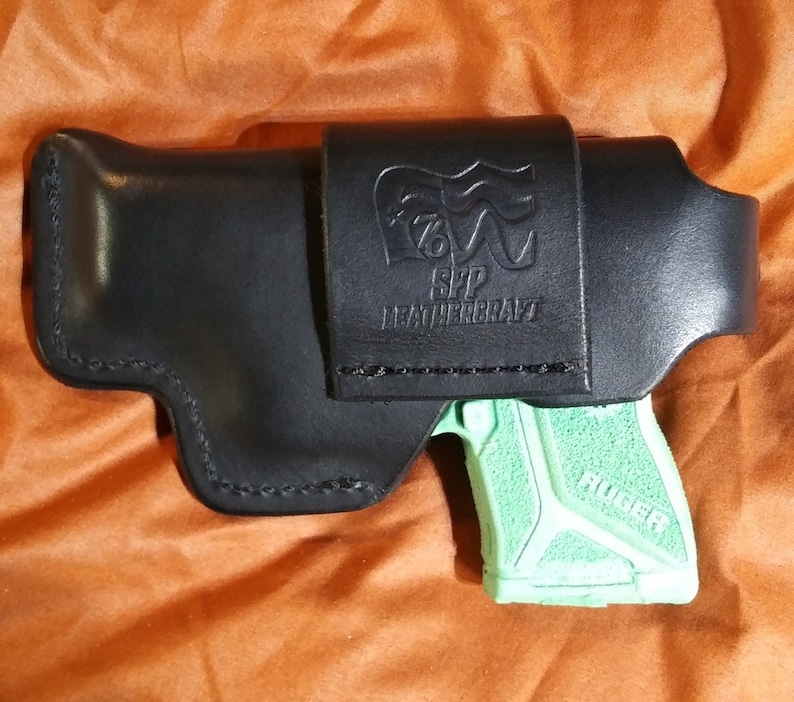 以下が含まれることがあります： 「SPP LEATHERCRAFT」の文字とロゴが入った黒い革製ホルスター。緑色の拳銃がホルスター内の一部に見えます。ホルスターはベルトに装着するように設計されています。背景はオレンジ色です。