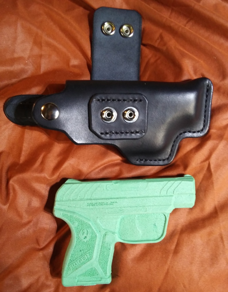 以下が含まれることがあります： 黒革製のホルスター。ベルト通しとスナップ留めが付いています。その下に、緑色の発泡スチロール製の拳銃レプリカがあります。ホルスターは銃器を収納するように設計されています。