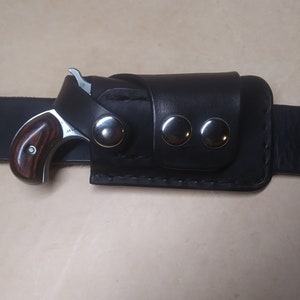 May include: A black leather holster with three silver snaps, designed to hold a small, wooden-handled handgun.
