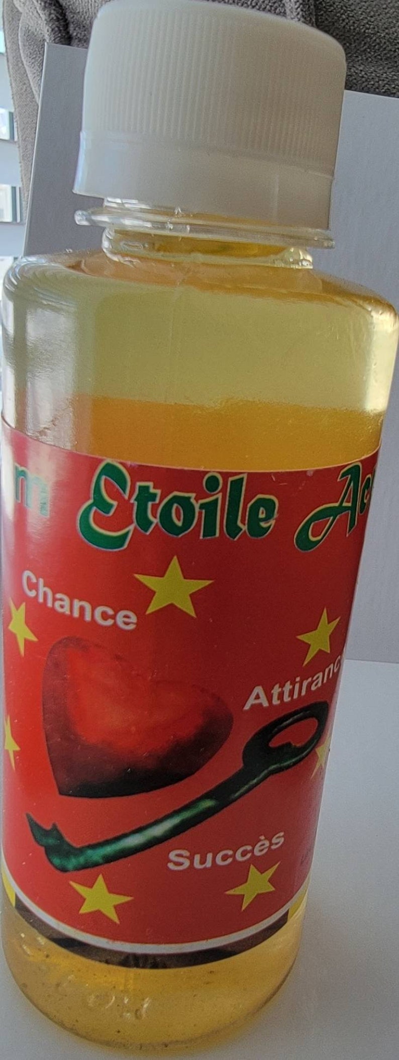 May include: A clear glass bottle with a white cap, filled with a yellow liquid. The bottle has a red label with green text and yellow stars. The label includes the words "Chance", "Attirance", and "Succ&egrave;s".