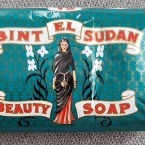 Può includere: Una saponetta blu turchese con lettere dorate che recitano "Bint El Sudan Beauty Soap". Il sapone presenta una donna con un vestito nero e rosso con fiori bianchi.