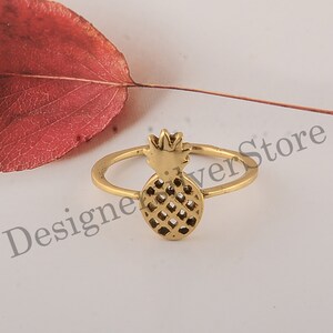 May include: A gold-toned ring with a pineapple design. The ring is on a white surface with a red leaf in the background.