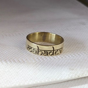 Pode incluir: Um anel dourado com a palavra "mahadev" gravada.
