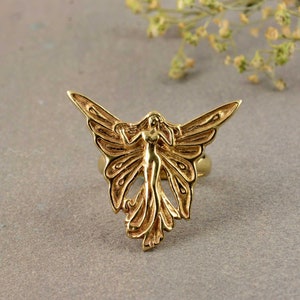 May include: A gold-toned ring featuring a detailed design of a fairy with wings spread wide. The fairy is depicted with a flowing gown and delicate features.