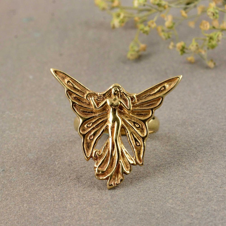 May include: A gold-toned ring featuring a detailed design of a fairy with wings spread wide. The fairy is depicted with a flowing gown and delicate features.