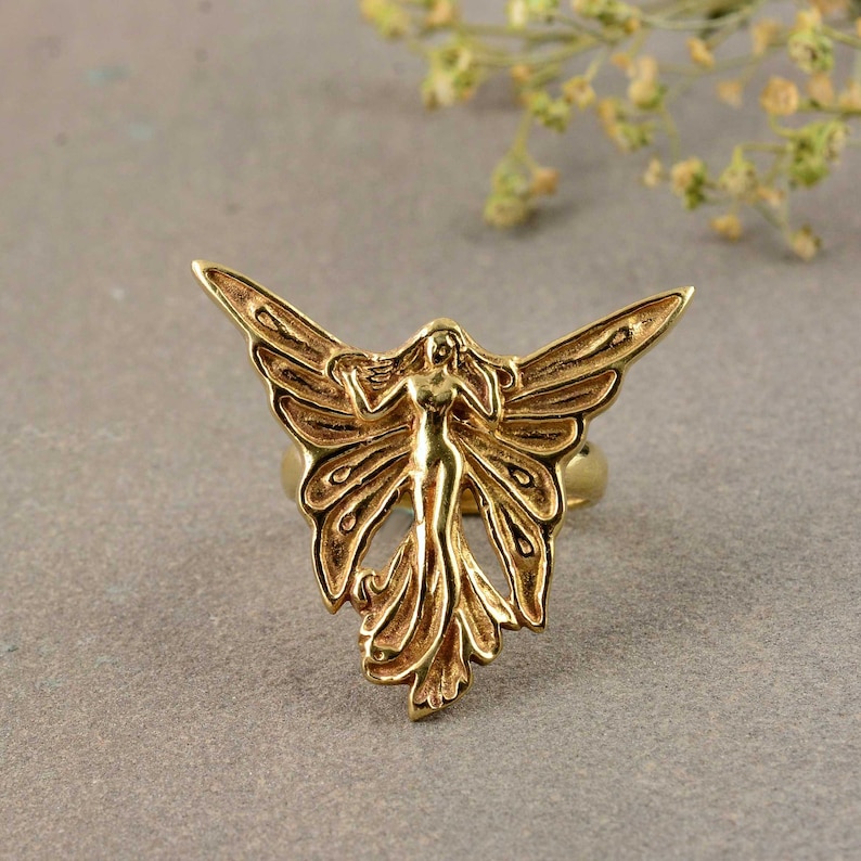 May include: A gold-toned ring featuring a detailed design of a fairy with wings spread wide. The fairy is depicted with a flowing gown and delicate features.