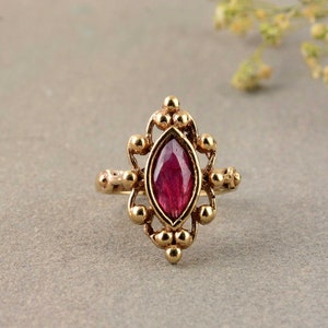 Może przedstawiać: Złoty pierścionek z ciemnobrązowym kamieniem szlachetnym osadzonym w delikatnym, ozdobnym wzorze.