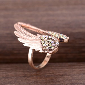 May include: A rose gold ring with a pair of wings adorned with iridescent green and clear rhinestones.