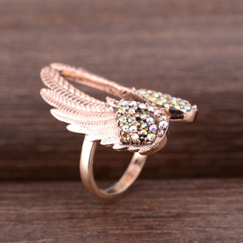 May include: A rose gold ring with a pair of wings adorned with iridescent green and clear rhinestones.