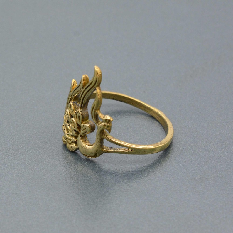 May include: A gold-toned ring with a peacock design. The peacock is facing to the right and has its wings spread out. The ring is made of metal and has a simple band.