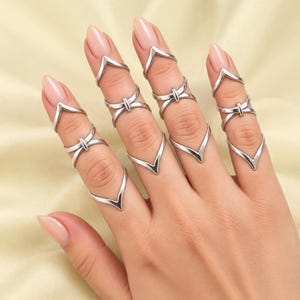 May include: A collection of silver-toned metal rings worn on the fingers. The set includes multiple V-shaped chevron rings and cross-over rings with a central vertical bar, designed to be worn as midi rings or stacked along the length of the fingers.
