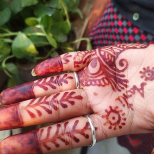May include: A hand with henna designs on the fingers and palm. The henna is a deep red colour and the designs are intricate. The hand is wearing two silver rings.