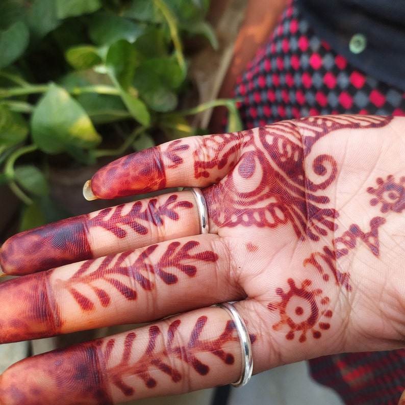 May include: A hand with henna designs on the fingers and palm. The henna is a deep red colour and the designs are intricate. The hand is wearing two silver rings.