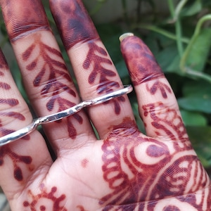 May include: A silver ring designed to be worn on multiple fingers. The ring is made of thick, textured metal and has a unique, asymmetrical shape.