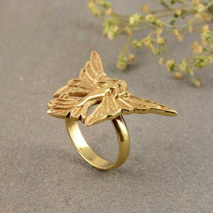 May include: A gold-toned ring with a detailed design of a fairy with wings spread out.