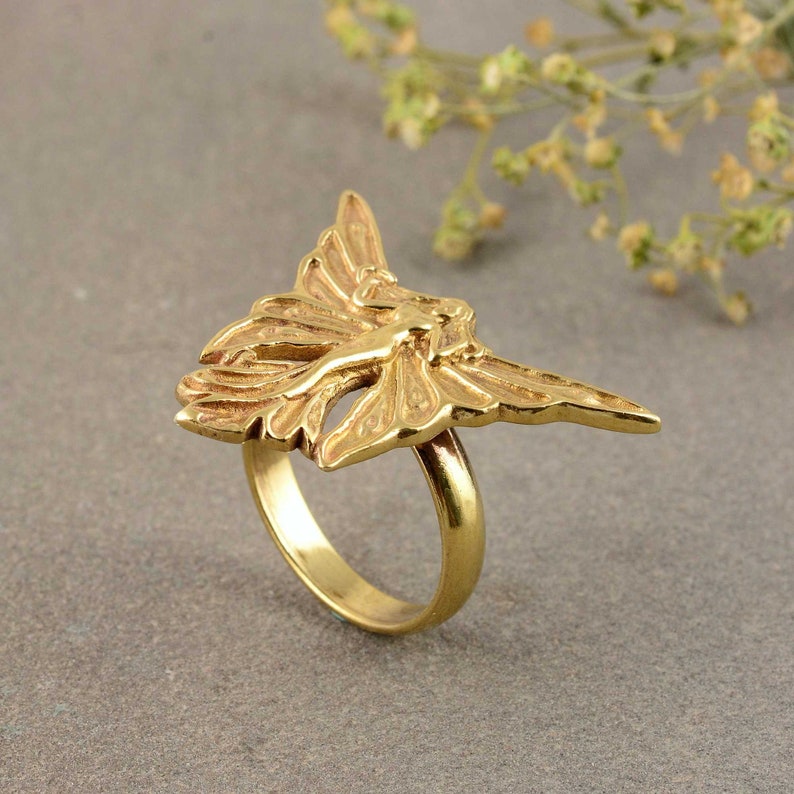 May include: A gold-toned ring with a detailed design of a fairy with wings spread out.