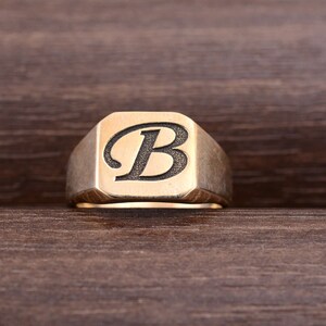 以下が含まれることがあります： 正方形の面に黒い文字「B」が刻まれた金のシグネットリングです。